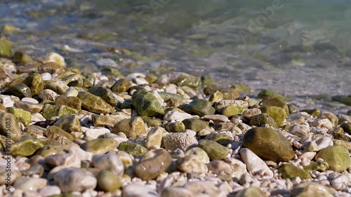 Close-up of pebbles covered in green algae on a beach, with gentle sea waves washing over them. The clear turquoise water adds to the tranquil ambiance.