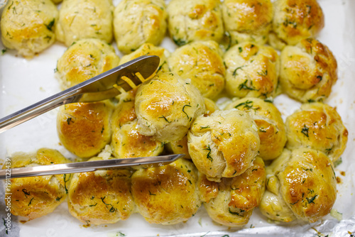 Photography Golden garlic herb dinner rolls lifted with metal tongs from baking tray, glossy