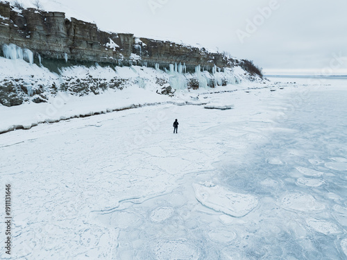 Aerial drone view of a lonely man walking on frozen sea ice near high limestone cliffs with icicles in Paldiski.