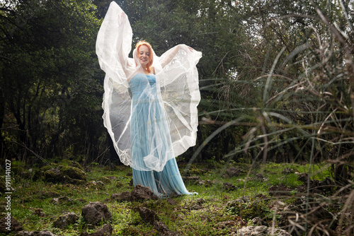 Ethereal woman in a light blue dress dancing with a white veil in the woods.