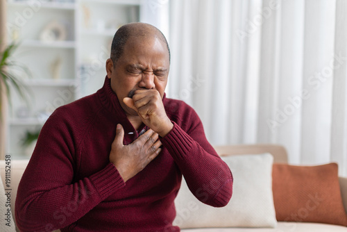 Fotografía Mature black man coughing with hand on chest while sitting on sofa at home