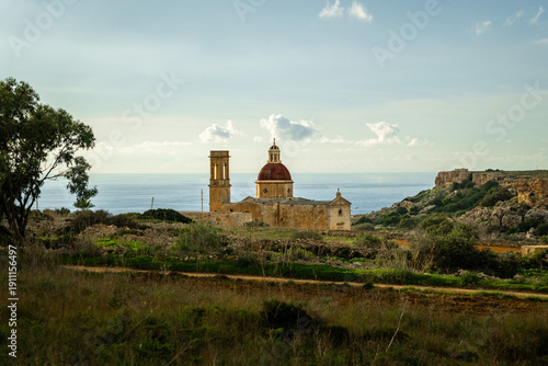 A historic Maltese church with a red dome stands above rocky hills near the coast—Nativity of the Virgin Mary glowing in soft Mediterranean light