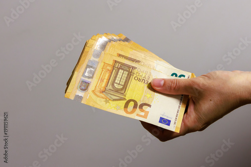A hand holds a thick stack of 50 euro banknotes against a light, neutral background. The banknotes are neatly stacked, and the distinctive euro design is clearly visible. 