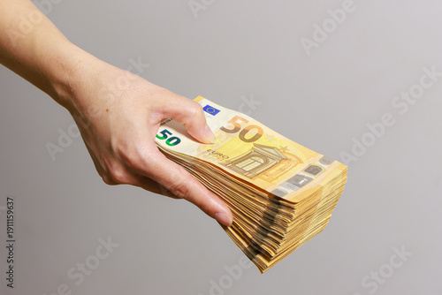 A hand holds a thick stack of 50 euro banknotes against a light, neutral background. The banknotes are neatly stacked, and the distinctive euro design is clearly visible. 