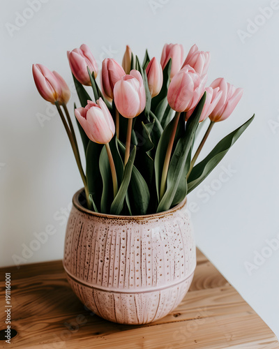 Pink tulips arranged in a decorative ceramic pot on a wooden surface, showcasing vibrant flowers with green leaves against a neutral background