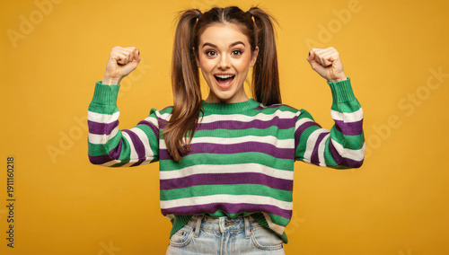 Vibrant Studio Portrait of a Joyful Young Woman in Striped Sweater Raising Fists in Victory Gesture Against Bright Yellow Background