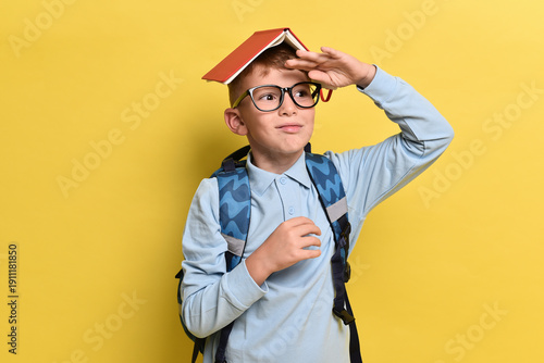Happy teenager boy holding backpack and book on yellow background . Red-haired boy with glasses