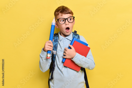 Happy teenager boy holding backpack and book on yellow background . Red-haired boy with glasses