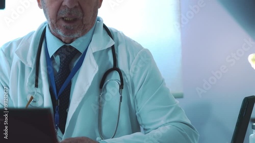 Elderly male physician with a stethoscope working at his desk in the medical clinic office