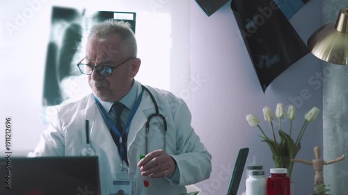 Medical doctor sitting at his desk, holding and looking at a test tube with a blood sample