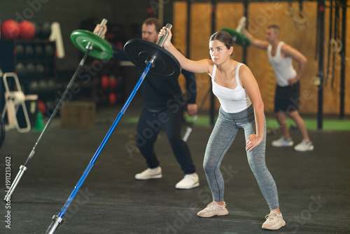 Tableau sur toile Determined sporty young girl doing one arm landmine press with barbell in high-intensity group training session at modern gym, focusing on shoulder and core muscles