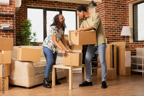 Tableau sur toile Smiling boyfriend and girlfriend carrying and arranging cardboard boxes in newly purchased apartment