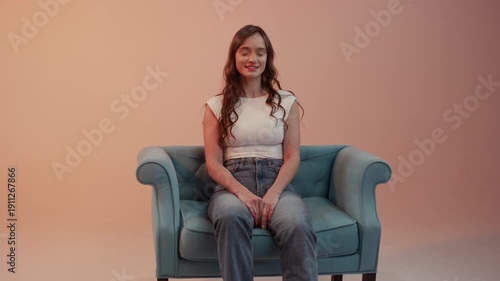 Studio shot, wide frame, of a young woman sitting centrally on a turquoise sofa. She is wearing jeans and a white top, smiling against a bright peach background. 4K.