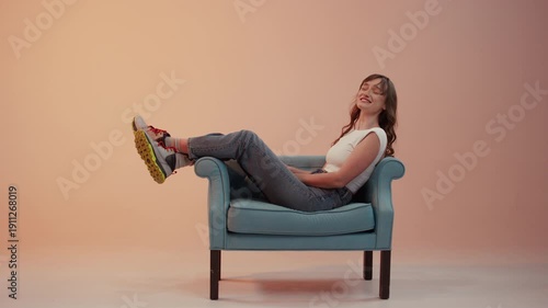 High quality, wide frame video of a young woman relaxing in a turquoise armchair. She is wearing jeans, sneakers and a white top, her legs raised against a peach background. 4K.