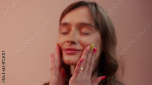 Close-up shot of a young Caucasian woman with colorful nails. She touches her cheeks with closed eyes, against a peach studio background. 4K.