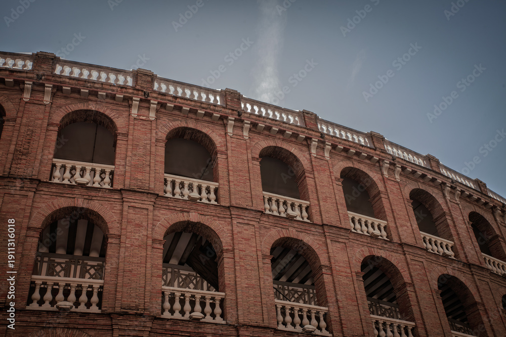 Naklejka premium Low angle view of Plaza de Toros de Valencia, a red brick bullring in Valencia, Spain, with arched galleries and white balustrades under a cloudy sky, highlighting a historic bullfighting landmark.