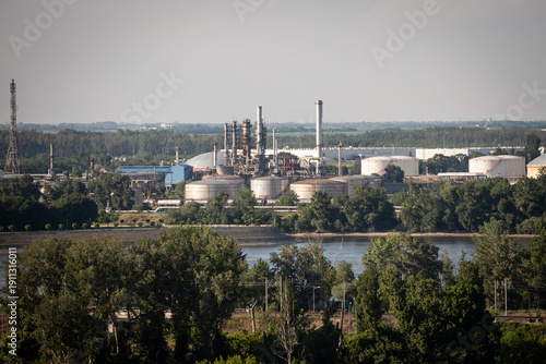Oil refinery complex in Serbia seen from a distance, with storage tanks, distillation towers, and smokestacks by a river. Industrial energy infrastructure linked to petrol production and policy.