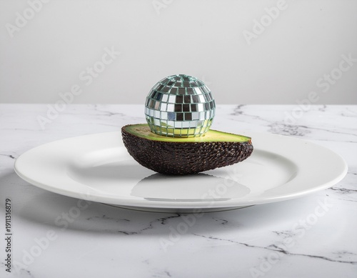 A surreal image of a small disco ball resting on a halved avocado on a white plate, set against a marble background.