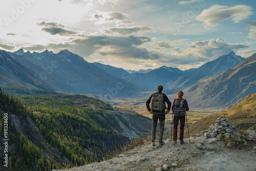 Couple of hikers with backpacks standing on mountain peak looking at vast valley, panoramic view of snow capped mountains and autumn forest in Mongolia, achievement and travel concept