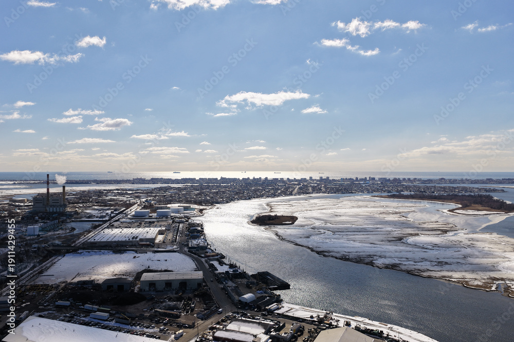 Obraz premium Aerial view of a cold winter port and power plant on long island sound, smoke pluming over partially frozen marshes, river channels and a distant new york cityscape under clear sky