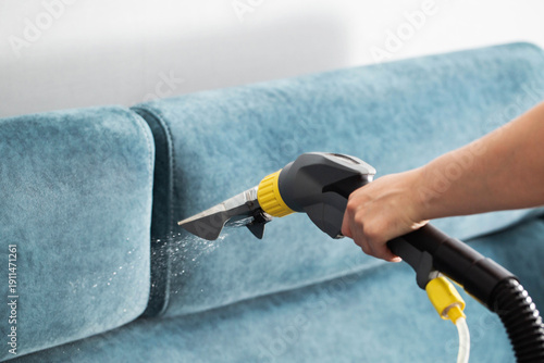 Professional deep cleaning of blue fabric sofa using powerful spray extraction machine by service worker hand, water droplets visible during process