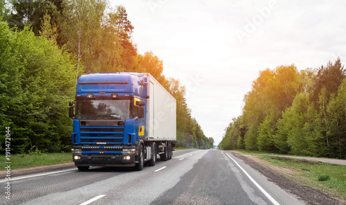 Blue heavy commercial truck with white semi-trailer transports cargo along open highway flanked by lush green forest under overcast sky during day