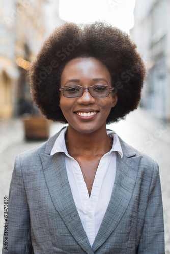 A professional Black woman with an afro hairstyle and glasses smiles confidently while wearing a grey suit jacket and white shirt outdoors on a city street