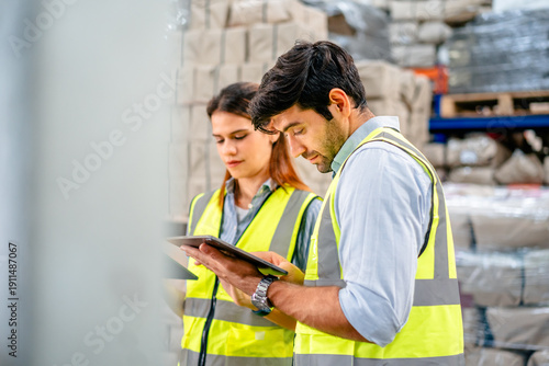 Diverse team of warehouse supervisors in safety vests using digital tablets to manage inventory and logistics in a large industrial printing facility storage area