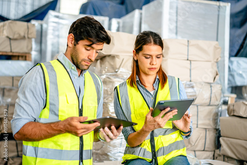 Diverse team of warehouse supervisors in safety vests using digital tablets to manage inventory and logistics in a large industrial printing facility storage area