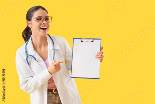 Young female medical student pointing at blank clipboard on yellow background