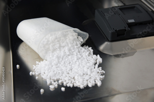 Foto Dishwasher salt pouring from translucent container onto metal surface, small white crystals used for water softening inside washing appliance