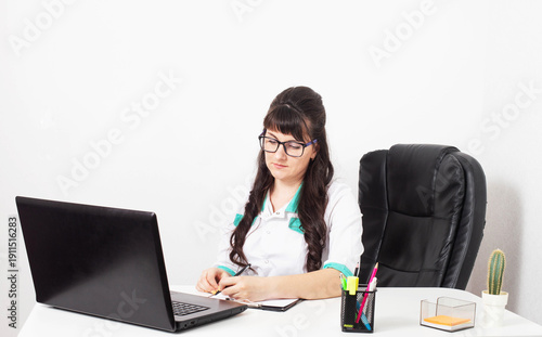 Female doctor or medical professional writing patient notes at office desk. Laptop computer, anatomical chart on wall behind. Medical work environment.