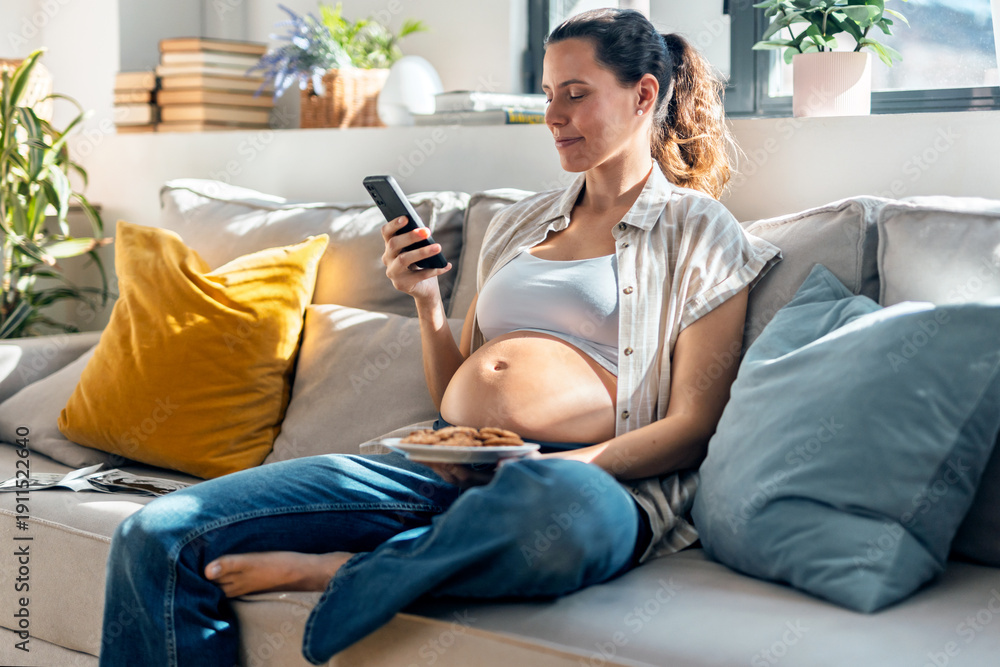 Obraz premium Pregnant woman using smartphone while eating a cookies sitting on the couch at home