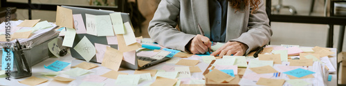 Caucasian woman working at cluttered desk covered with sticky notes, writing on paper beside open laptop, demonstrating busy business planning and organization process