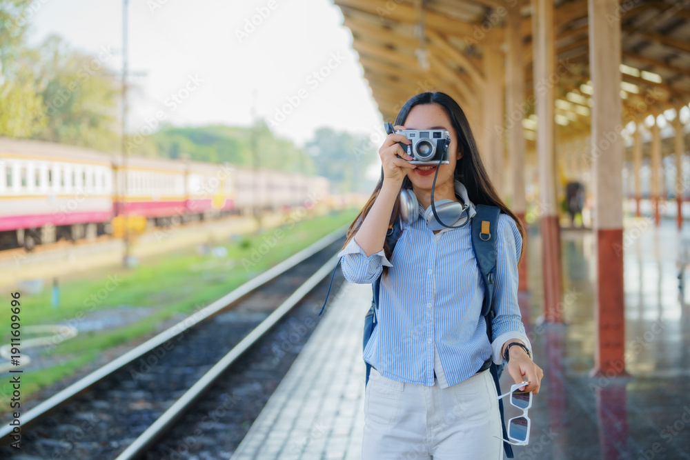 Fototapeta premium A girl smiles, holding a film camera. Young Asian woman traveler with a camera taking pictures on a train platform. Adventure, hangout, travel, holiday, relaxation, photography concept.