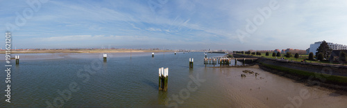 A tranquil coastal scene featuring a shallow river estuary, wooden piers, and a sandy shoreline under a bright blue sky.