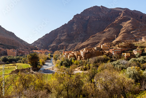 A village of earthen buildings nestled between mountains, with a river flowing through it. Marrakech region,Morocco