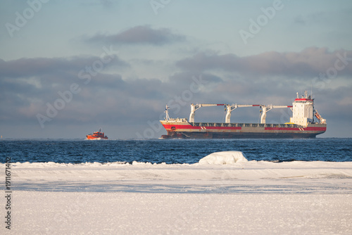 Large cargo ship and pilot boat navigating through cold blue waters of a frozen sea near snowy arctic winter coast
