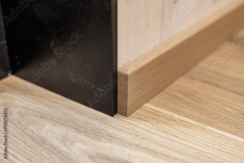 Fotografi Close-up of a wooden baseboard meeting a light wood-paneled wall and floor, adjacent to a black metal frame