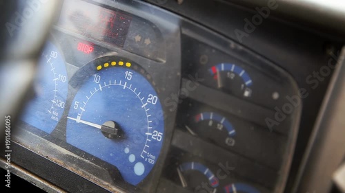 Instrument panel of vehicle showing speedometer, tachometer, and fuel gauge. Red stop warning light illuminated. Surface of dashboard appears dusty. Blue and black color scheme prominent.