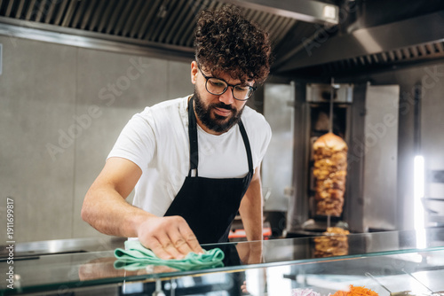 Billede på lærred Male food vendor with curly hair wearing glasses cleans glass display case in a