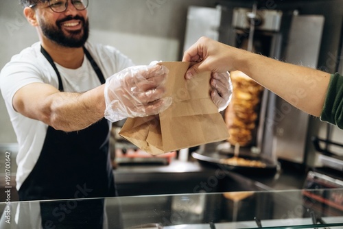 Fényképezés Male food vendor in black apron hands over a brown paper bag to a customer at a