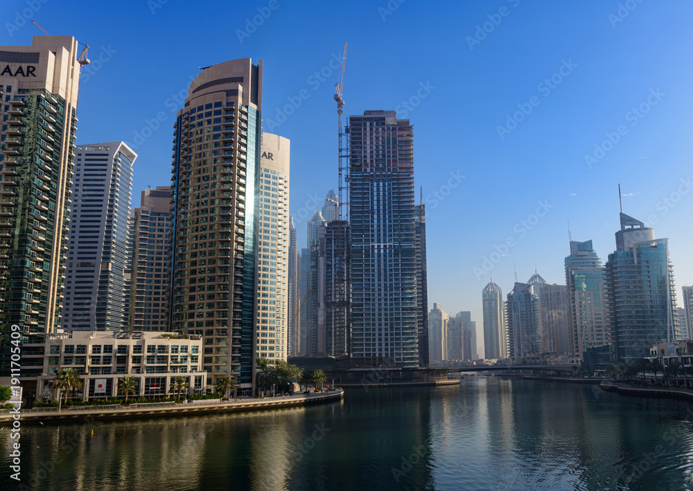 Obraz premium Dubai Marina skyline with modern skyscrapers and waterfront reflections under clear blue sky, luxury urban architecture in UAE.