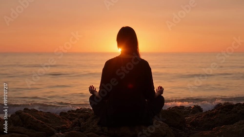 Woman meditating on rocky beach at serene sunset with warm orange glow in peaceful silhouette for wellness advertising