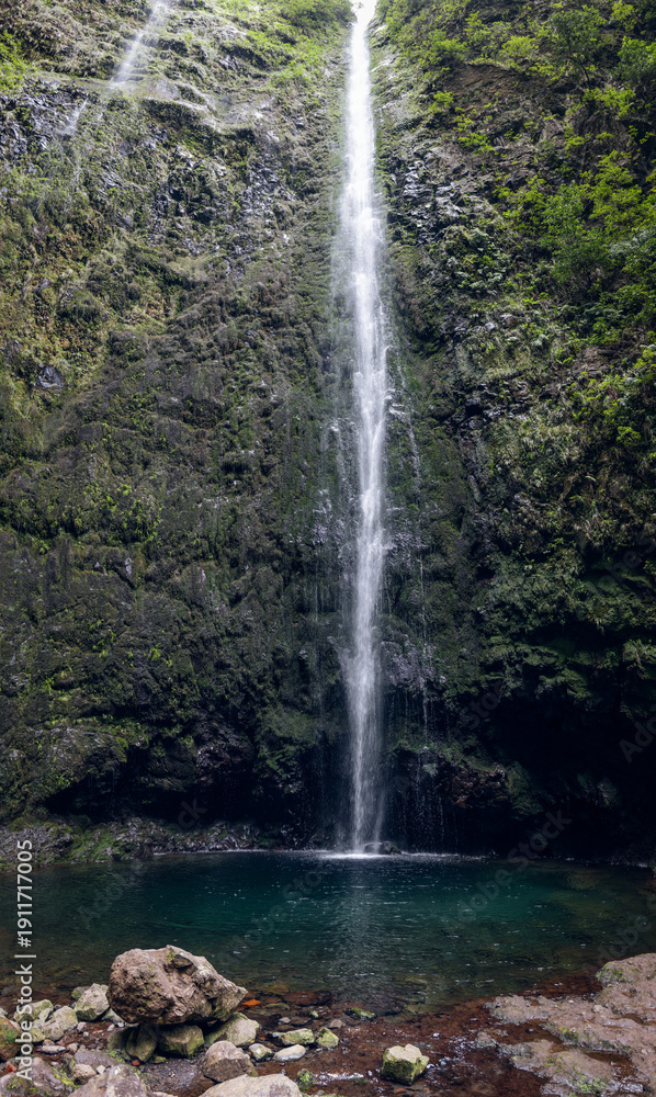 Fototapeta premium Caldeirao Verde Waterfall at trail end on route PR9, thin white plunge drops into an emerald pool beneath mossy basalt walls with ferns and soft mist (vertical panoramic photo)