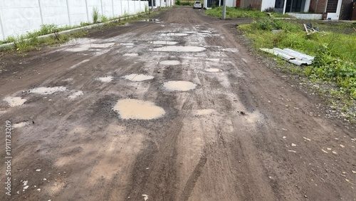 Konstfotografi Muddy unpaved road with numerous water-filled potholes, tire tracks, and green grass on the sides, stretching towards a distant car and residential buildings
