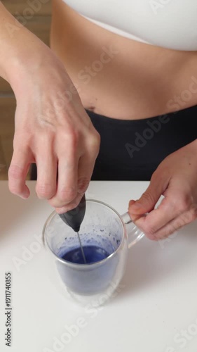 Woman frothing blue matcha latte in clear glass cup at home
