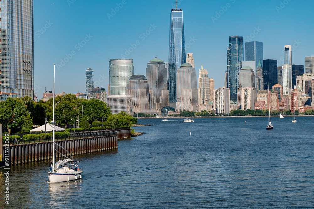 Obraz premium Sailing boat on Hudson River with Lower Manhattan skyline. White sailboat navigating water near a park with skyscrapers in background.