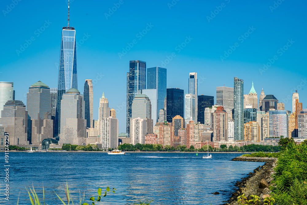Obraz premium Lower Manhattan skyline from the shore with green foliage. Skyscrapers and One World Trade Center viewed over water with rocky shoreline and grass.