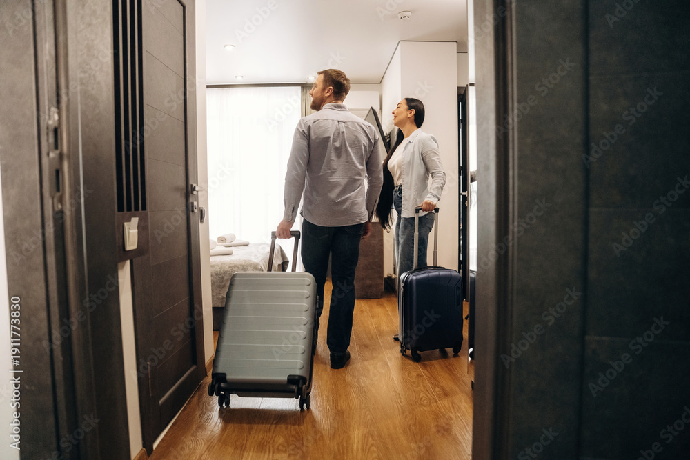 Fototapeta premium Couple entering hotel room carrying luggage, modern interior design with wooden flooring, large window providing natural light, and a cozy atmosphere visible in the background